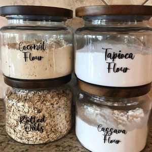 May include: Four clear glass jars with wooden lids, each filled with a different ingredient. The jars are labeled "Coconut Flour", "Tapioca Flour", "Rolled Oats", and "Cassava Flour". The jars are stacked on top of each other.
