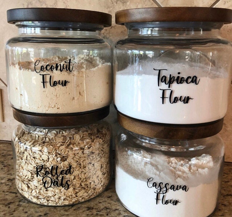 May include: Four clear glass jars with wooden lids, each filled with a different ingredient. The jars are labeled "Coconut Flour", "Tapioca Flour", "Rolled Oats", and "Cassava Flour". The jars are stacked on top of each other.