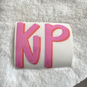 May include: A pink and orange sticker with the letters "KP" in a bold, layered design. The letters are a bright pink with an orange shadow, set against a white background. The sticker is placed on a fluffy, white surface.