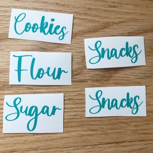 May include: Five white rectangular labels with teal cursive text. The labels read "Cookies", "Flour", "Sugar", and two that read "Snacks". The labels are arranged on a light brown wooden surface.