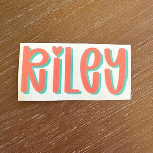 May include: A rectangular sticker with the name "Riley" in coral-colored, bubble-style letters with a teal outline. A small heart replaces the dot above the "i". The sticker is on a wooden surface.