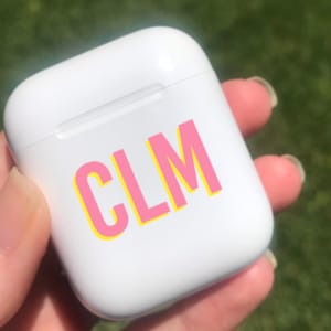 Op de afbeelding: Witte draadloze oplaadcase met een roze en gele sticker met de tekst "CLM" erop.