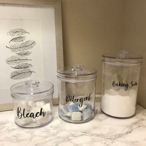 May include: Three clear, lidded canisters labeled "Bleach", "Detergent", and "Baking Soda". The "Bleach" canister contains white pods, the "Detergent" canister has blue and white pods, and the "Baking Soda" canister is filled with white powder. A framed feather art piece is in the background.