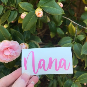 May include: A white sticker with the word "Nana" written in pink cursive lettering. The sticker is being held in front of a green bush with pink flower buds.