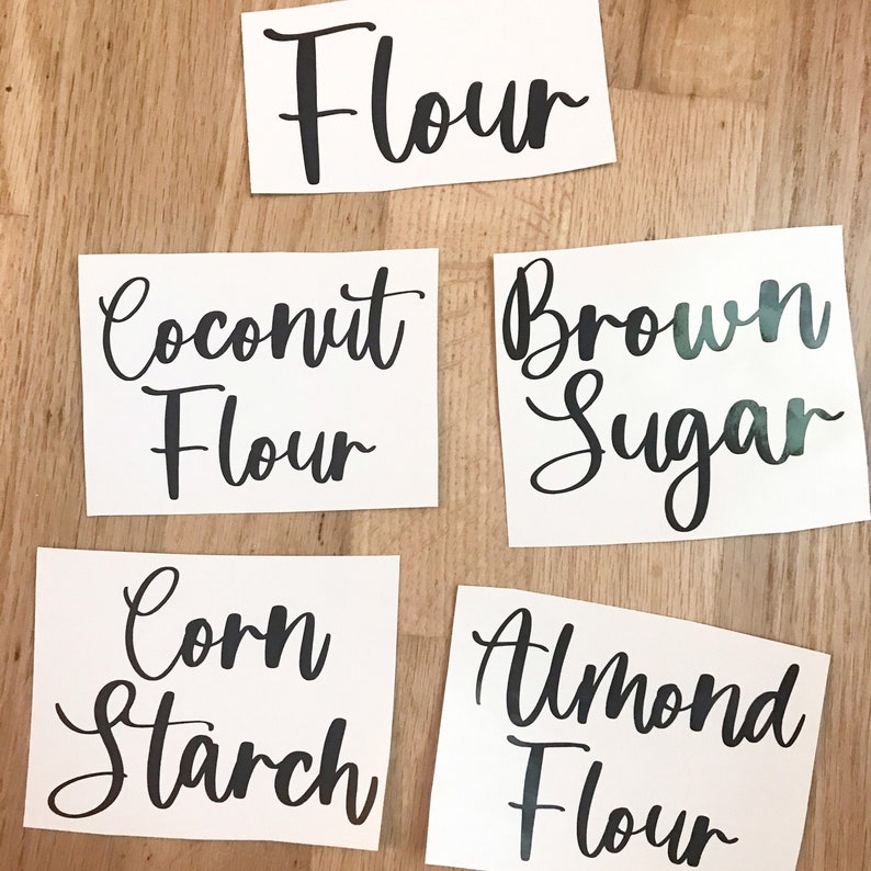 May include: Five white rectangular labels with black cursive text on a light brown wooden surface. The labels read "Flour", "Coconut Flour", "Brown Sugar", "Corn Starch", and "Almond Flour".