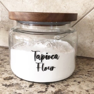 May include: A clear glass jar filled with white tapioca flour, topped with a wooden lid. The words "Tapioca Flour" are written in black script on the front of the jar. The jar is sitting on a speckled countertop.