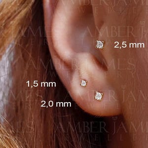 May include: Close-up of an ear with three small, gold-colored stud earrings, each with a clear, square-cut stone. The earrings are 1.5 mm, 2.0 mm, and 2.5 mm in size. The text "2,5 mm", "1,5 mm", and "2,0 mm" is visible.