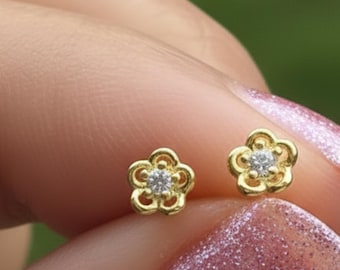 Könnte beinhalten: Zwei kleine goldene Ohrstecker in Form von fünfblättrigen Blumen, jeweils mit einem einzelnen klaren, runden Edelstein in der Mitte. Die Ohrringe haben ein poliertes Gold-Finish und ein filigranes durchbrochenes Design. Text AMBER JAMES JEWELLERY.