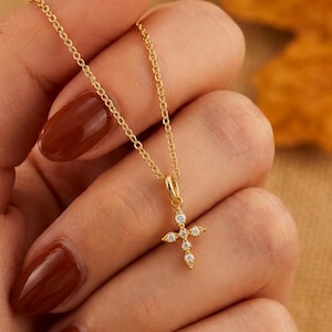 May include: A delicate gold chain necklace featuring a small cross pendant adorned with tiny clear stones. The cross is centred on the chain, which is held up to display the jewellery piece clearly. The text AMBER JAMES JEWELLERY is visible.