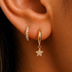 May include: A gold-toned ear piercing set consisting of a small star-shaped stud with a central clear stone in the upper cartilage and a gold hoop earring in the lobe featuring a dangling star charm encrusted with multiple small clear stones.