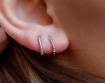 Pode incluir: Dois pequenos brincos de argola em prata com uma superfície texturizada de contas. Os brincos são usados em dois furos adjacentes no lóbulo inferior da orelha, apresentando um acabamento metálico polido que reflete a luz com um padrão sutil.