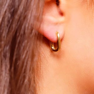 Può includere: Un piccolo orecchino a cerchio color oro con una distinta forma rettangolare. L'orecchino presenta angoli arrotondati e una superficie metallica liscia e riflettente, fissato saldamente al lobo dell'orecchio, mostrando un design semplice e pulito.