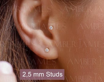 May include: Three small, round, clear stud earrings are positioned in the earlobe and cartilage. Each stud features a single, brilliant-cut stone set in a metallic base. A text overlay on the image clearly indicates the size as 2.5 mm Studs.