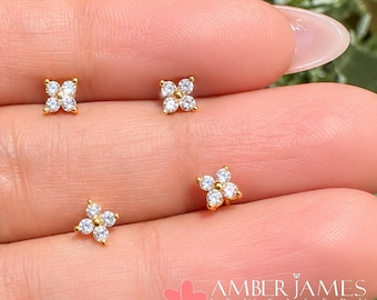 Puede incluir: Cuatro pendientes de botón dorados, cada uno con un diseño de flor de cuatro pétalos engastado con piedras de circonita cúbica transparentes y brillantes. Los pendientes están dispuestos en forma cuadrada. El texto AMBER JAMES JEWELLERY aparece abajo.