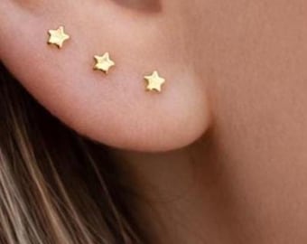 Puede incluir: Tres pequeños pendientes de botón con forma de estrella dorada, dispuestos en una fila horizontal a lo largo del cartílago inferior de la oreja. Cada pendiente presenta un acabado metálico pulido y una silueta clásica de cinco puntas.