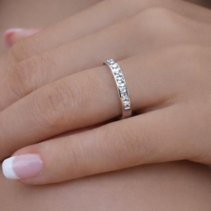 May include: A silver ring set with a line of small, clear gemstones. The ring is on a person's finger, with the hand and arm visible. The ring has a simple band design, suitable for everyday wear or as a wedding band.