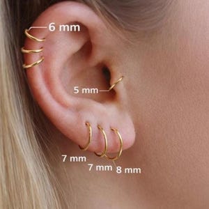May include: An ear displaying several gold hoop earrings of varying sizes. The labels indicate sizes of 6 mm, 5 mm, 7 mm, and 8 mm. The earrings offer a minimalist aesthetic, highlighting a simple jewellery design.
