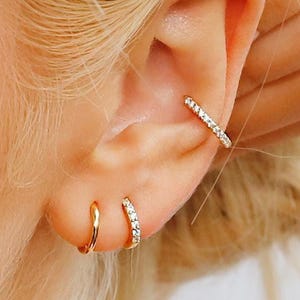 May include: Close-up of an ear adorned with three earrings. One is a gold-colored ear cuff with small, clear stones. The other two are gold-colored hoops, one plain and one with clear stones. The text at the bottom reads "Sterling silver pave hoops For plain hoops find link below."