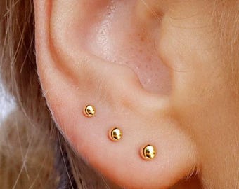May include: Three small, polished gold ball stud earrings are arranged in a row along the earlobe. Each earring features a smooth, spherical design with a reflective metallic finish, creating a simple and uniform look across the lower ear.