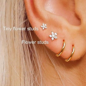 May include: Two small flower-shaped stud earrings with clear stones and two plain gold hoop earrings arranged in a vertical row on an earlobe. The jewellery pieces are labelled with the text Tiny flower studs and Flower studs.