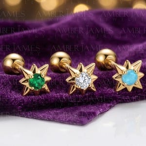 Puede incluir: Pendientes de estrella dorados con gemas verdes, blancas y turquesas. Los pendientes están presentados sobre una superficie de terciopelo morado. Cada pendiente tiene un pequeño perno redondo.