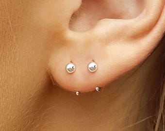May include: Close-up of an ear with three pairs of small, silver stud earrings. A silver hoop earring with a small ball design is worn in the upper ear. The earrings have a simple, minimalist design.
