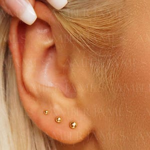 May include: Three small, polished gold ball stud earrings arranged in a row along the lower earlobe. Each earring features a smooth, spherical design with a reflective metallic finish, creating a simple and uniform look across the ear piercing.