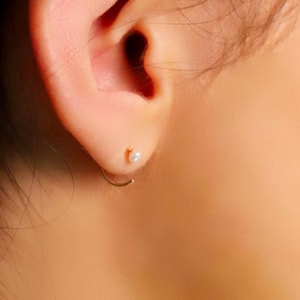 Op de afbeelding: Een delicate gouden oorring met een kleine, ronde witte parel als accent. De oorbel wordt in de oorlel gedragen en toont een dun, minimalistisch gouden draadontwerp dat zachtjes buigt. De tekst AMBER JAMES JEWELLERY is onderaan zichtbaar.
