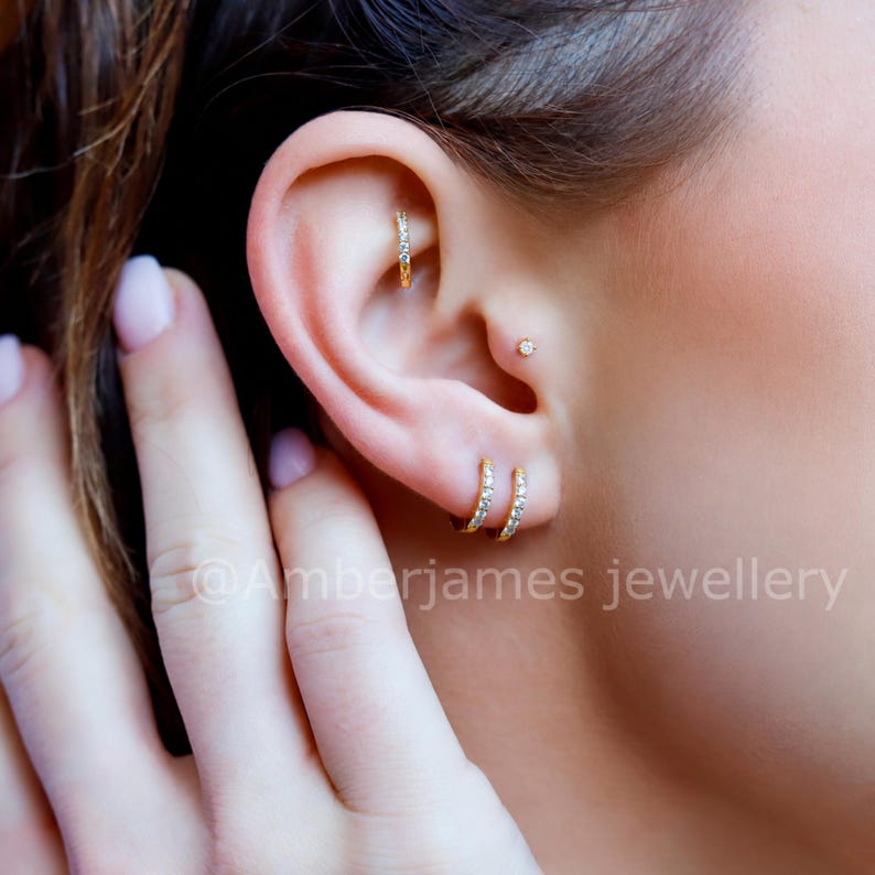 Puede incluir: Pendientes de aro dorados y un peque&ntilde;o pendiente de bot&oacute;n con piedras claras y brillantes. Las joyas se llevan en el l&oacute;bulo y el cart&iacute;lago. El texto @Amberjames jewellery es visible en la parte inferior de la imagen.