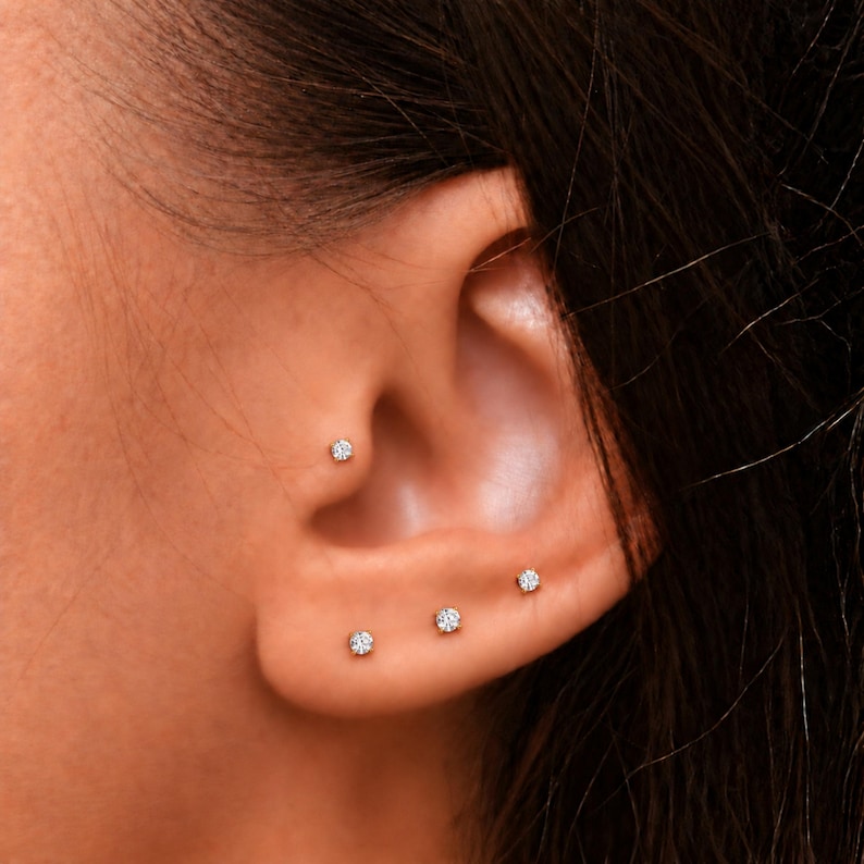 May include: A set of four small, round, clear stud earrings worn in the ear. One stud is placed in the upper cartilage area, while the other three studs are arranged in a line along the earlobe. Each earring features a single, brilliant-cut stone.
