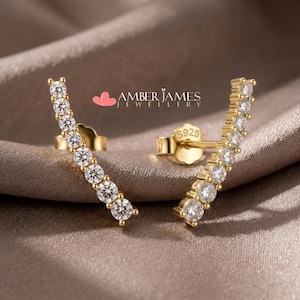 Può includere: Un paio di orecchini a lobo dorati con una fila di pietre preziose rotonde e trasparenti. Gli orecchini sono appoggiati su un tessuto dai toni neutri. Il testo "AMBER JAMES JEWELLERY" è visibile.