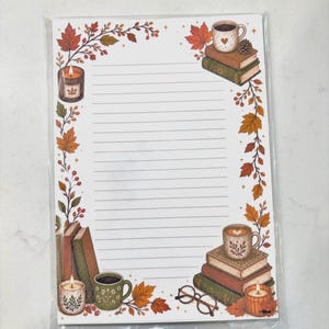 May include: A rectangular sheet of lined stationery featuring a decorative border of autumn leaves, berries, and small stars. The design includes illustrations of stacked books, mugs of coffee, lit candles, and eyeglasses in the corners.