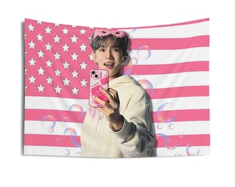 May include: A pink and white American flag tapestry with a person holding a pink phone case. The person is wearing a pink eye mask and a cream-colored hoodie. Bubbles float in the air in front of the tapestry.