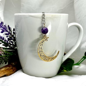 May include: Gold crescent moon tea infuser with a purple amethyst bead. The infuser is hanging from a silver chain and is in a white teacup.