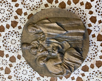 May include: A bronze-colored, circular relief sculpture depicting several figures in draped garments. The figures are arranged in a scene, with some kneeling and others standing. The sculpture is set against a white lace doily.