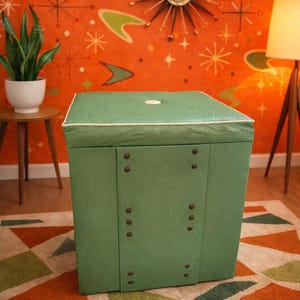 May include: Green square ottoman with a white trim and button detail. The sides are adorned with decorative rivets. The ottoman sits on a geometric patterned rug, against an orange wall with mid-century modern decor.