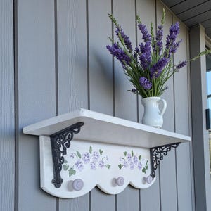May include: A white wooden shelf with a scalloped edge, adorned with purple floral designs and three lavender knobs. It features black metal brackets and is mounted on a gray wall. A white pitcher holds purple lavender sprigs.