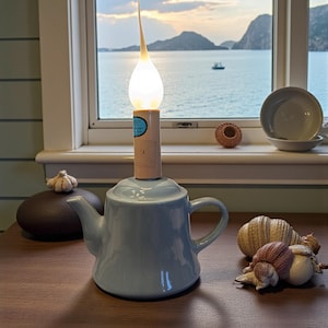May include: A light blue ceramic teapot with a unique lamp fixture. The lamp has a long, slender, curved stem and a teardrop-shaped light. The lamp is placed on top of the teapot. The background shows a scenic view of the sea and mountains.