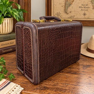 May include: A vintage-style dark brown suitcase featuring a textured faux crocodile skin pattern. The rectangular case includes a sturdy top handle, gold-toned metal latches, and reinforced edges with visible stitching along the side panels.