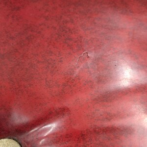 Puede incluir: Primer plano de una superficie roja texturizada con una peque&ntilde;a hendidura. La superficie parece estar hecha de un material brillante, con algunas manchas oscuras dispersas. Una porci&oacute;n de un objeto redondo de color claro es visible en la esquina inferior izquierda.