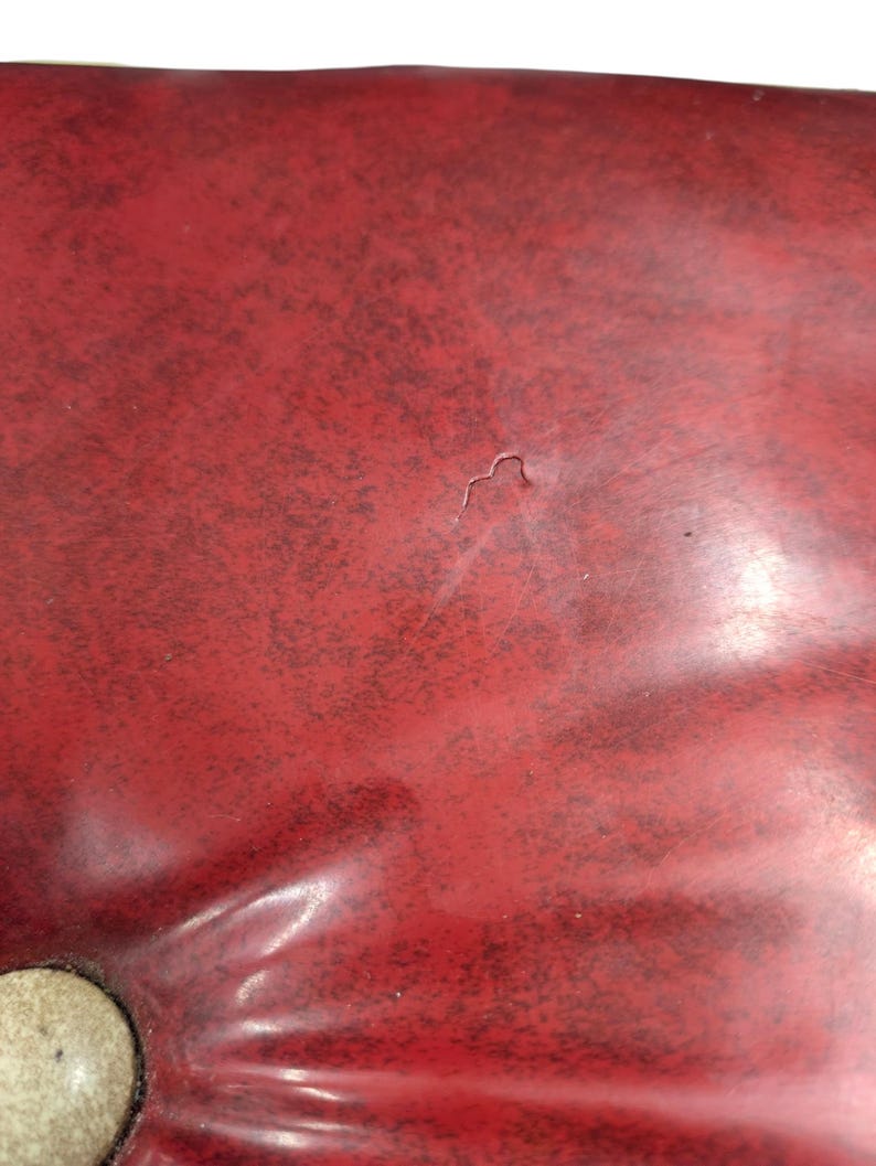 Puede incluir: Primer plano de una superficie roja texturizada con una peque&ntilde;a hendidura. La superficie parece estar hecha de un material brillante, con algunas manchas oscuras dispersas. Una porci&oacute;n de un objeto redondo de color claro es visible en la esquina inferior izquierda.