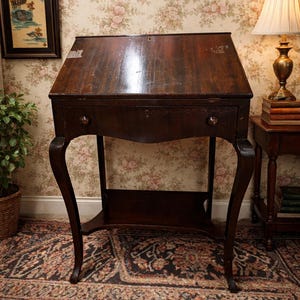 May include: A dark brown antique writing desk with a slanted top and a single drawer. The desk has curved legs and a small shelf. The background features floral wallpaper and a patterned rug. A lamp and framed artwork are also visible.