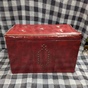 Puede incluir: Un otom&aacute;n de almacenamiento rojo vintage con una parte superior acolchada y un dise&ntilde;o decorativo con tachuelas en la parte delantera. El otom&aacute;n tiene forma rectangular y un ribete blanco alrededor del borde superior.