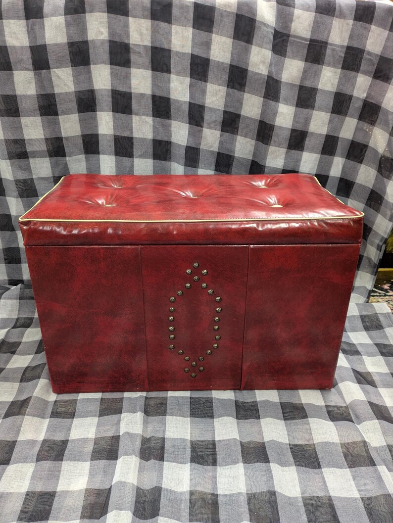 Puede incluir: Un otom&aacute;n de almacenamiento rojo vintage con una parte superior acolchada y un dise&ntilde;o decorativo con tachuelas en la parte delantera. El otom&aacute;n tiene forma rectangular y un ribete blanco alrededor del borde superior.