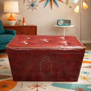 Puede incluir: Un otom&aacute;n rojo, acolchado con un ribete dorado y un dise&ntilde;o decorativo con tachuelas. El otom&aacute;n de estilo moderno de mediados de siglo tiene forma cuadrada y se asienta sobre una alfombra estampada colorida. La habitaci&oacute;n tiene una est&eacute;tica retro.