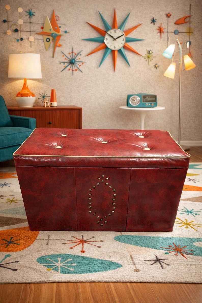 Puede incluir: Un otom&aacute;n rojo, acolchado con un ribete dorado y un dise&ntilde;o decorativo con tachuelas. El otom&aacute;n de estilo moderno de mediados de siglo tiene forma cuadrada y se asienta sobre una alfombra estampada colorida. La habitaci&oacute;n tiene una est&eacute;tica retro.