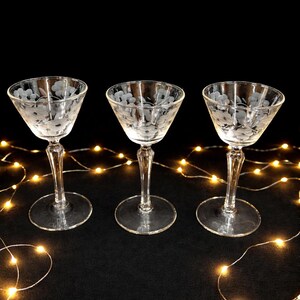 May include: Three clear, stemmed cocktail glasses with etched floral designs. The glasses have a classic coupe shape and sit on a black surface, with a string of warm white fairy lights in the background.