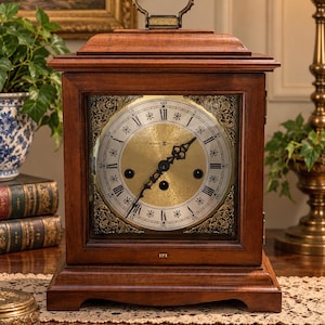 May include: A wooden Howard Miller mantel clock with a square frame, a gold-toned circular dial featuring Roman numerals, and ornate corner filigree. The clock has black hands, a top carrying handle, and the letters HM engraved on the base.
