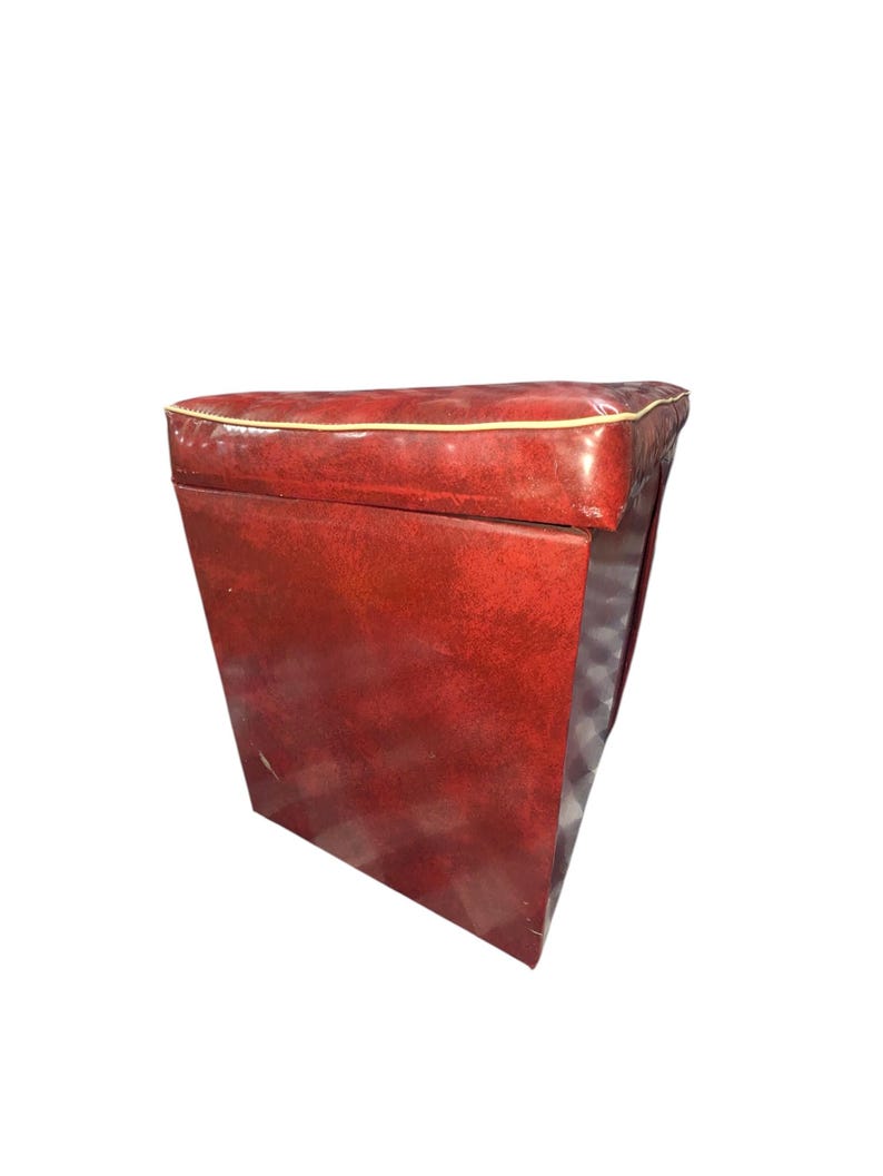 Puede incluir: Un otom&aacute;n rectangular rojo con una parte superior acolchada y un ribete dorado. Los lados son de color rojo liso y el interior es visible. El otom&aacute;n es de estilo vintage.