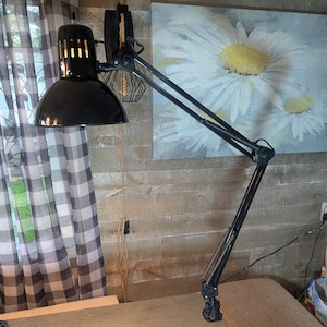 May include: A black adjustable desk lamp with a wide, rounded shade and a multi-jointed swing arm. The lamp features a clamp base for attachment to a surface and includes a series of vertical ventilation slots near the top of the lamp head.